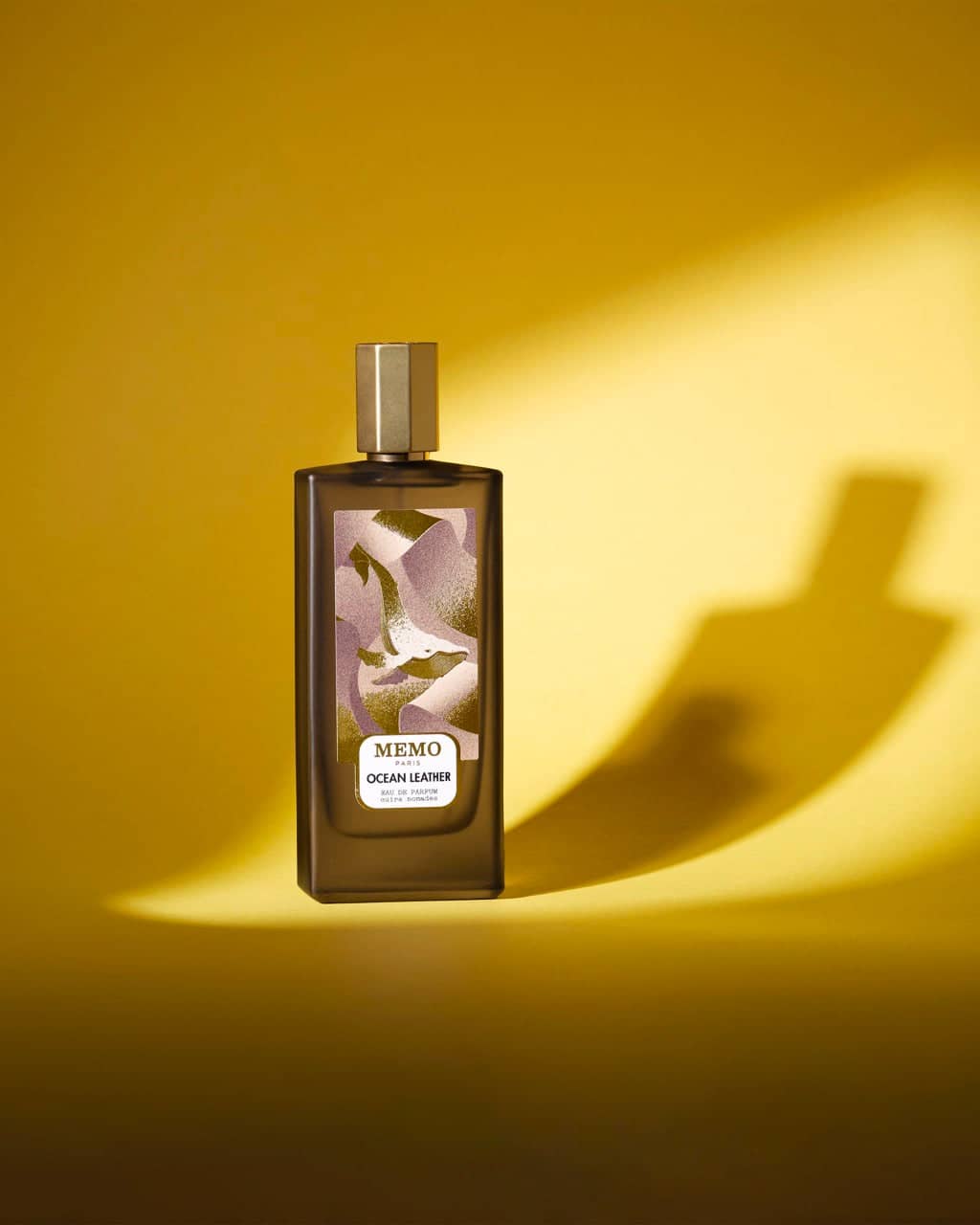 A sleek, dark brown rectangular perfume bottle with a gold cap and an abstract lavender-and-white label featuring the ’memo paris ocean leather’ branding.
